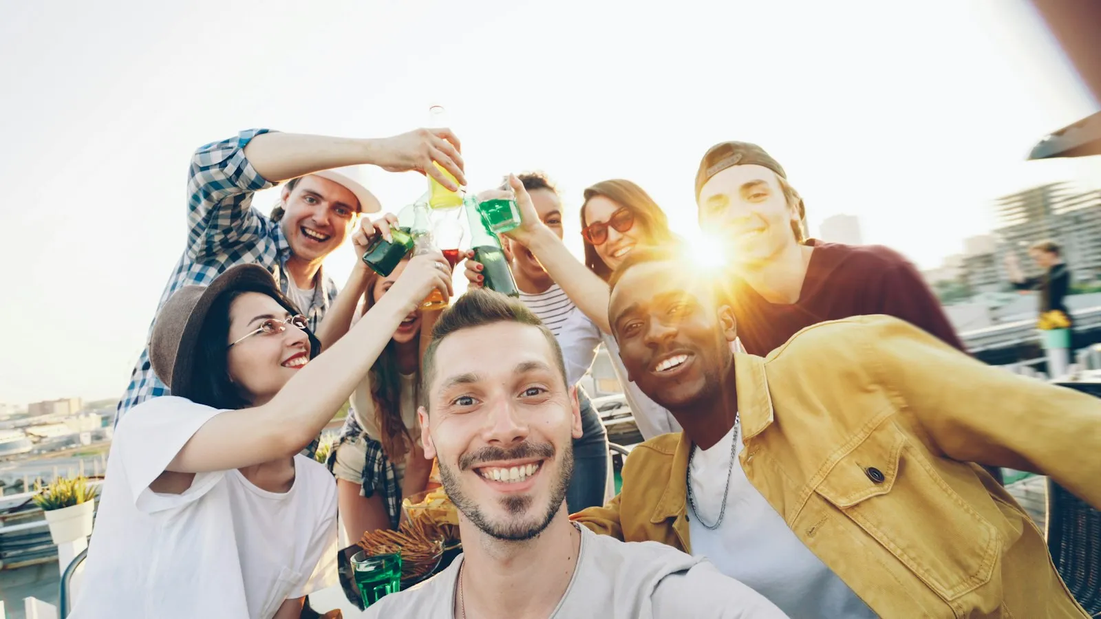 A laughing group of friends raising bottles and glasses together at a sunny rooftop gathering