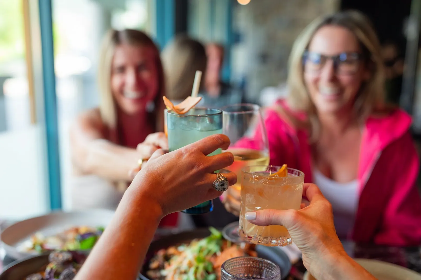 Two friends smiling and clinking cocktails over dinner at a bright social gathering
