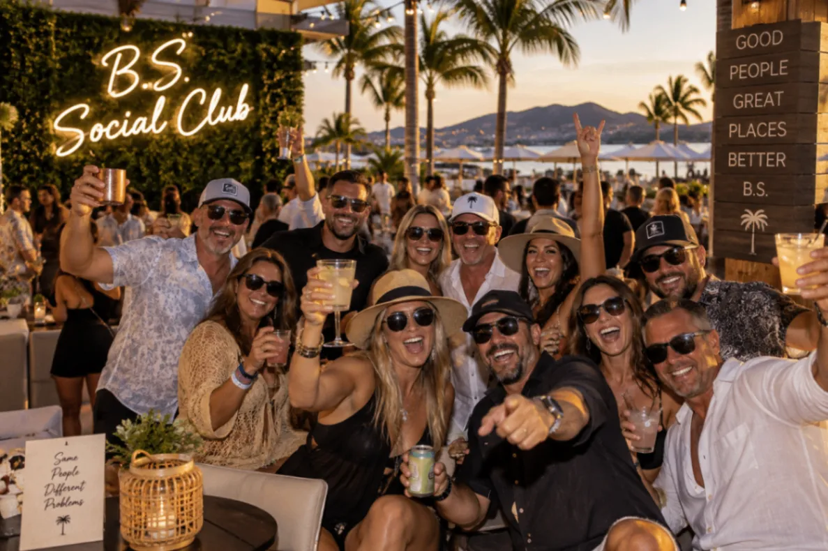 A bright B.S. Social Club group gathering with friends raising cocktails among palm trees at a waterfront destination